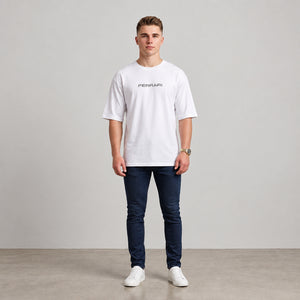 A man wearing a white short-sleeve t-shirt with a black 'FERRARI' logo on the chest, paired with dark blue jeans and white sneakers, standing against a neutral studio background l AH Urban Wear