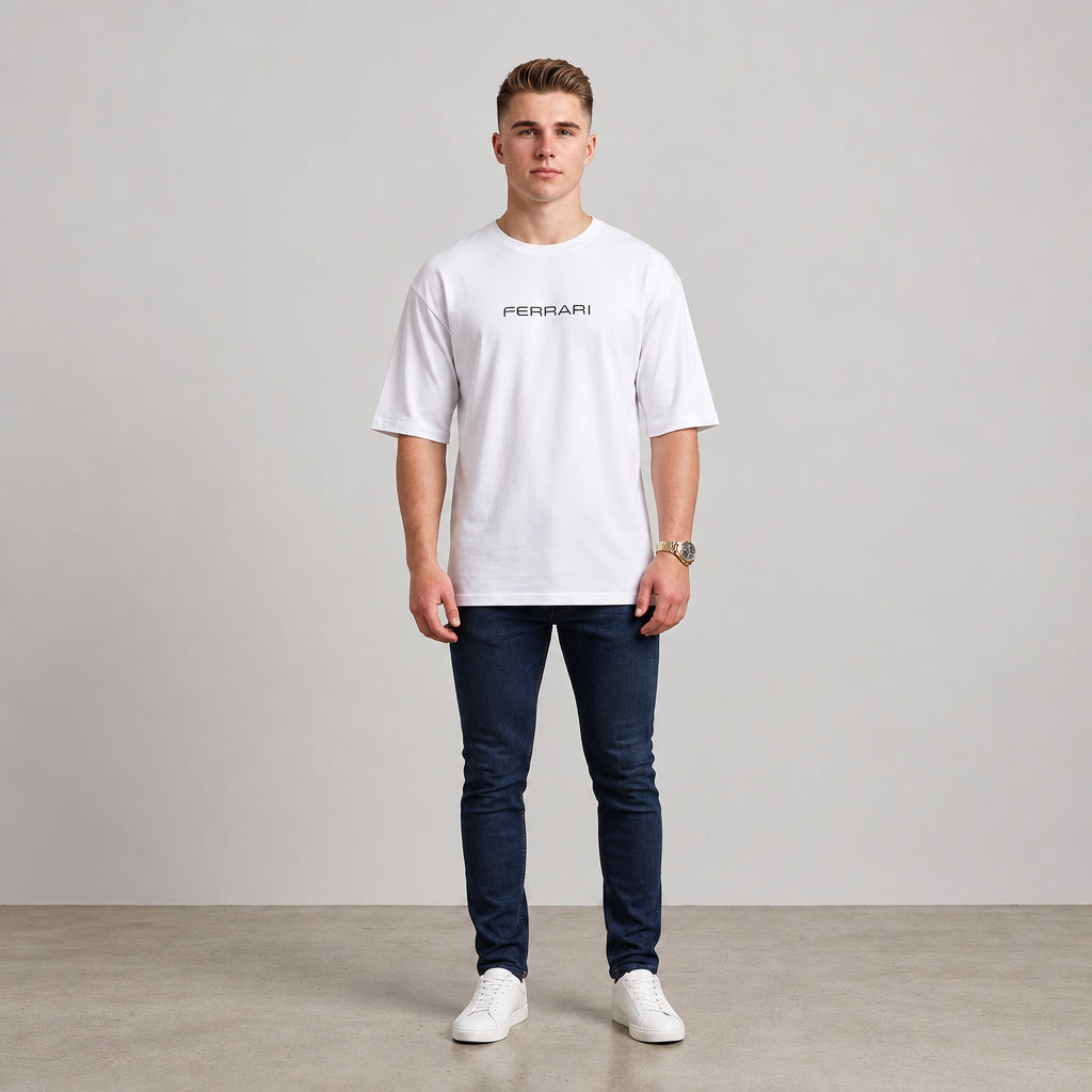 A man wearing a white short-sleeve t-shirt with a black 'FERRARI' logo on the chest, paired with dark blue jeans and white sneakers, standing against a neutral studio background l AH Urban Wear
