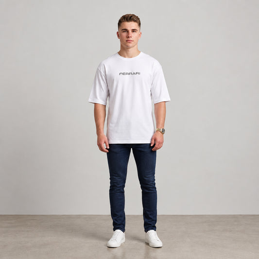 A man wearing a white short-sleeve t-shirt with a black 'FERRARI' logo on the chest, paired with dark blue jeans and white sneakers, standing against a neutral studio background l AH Urban Wear