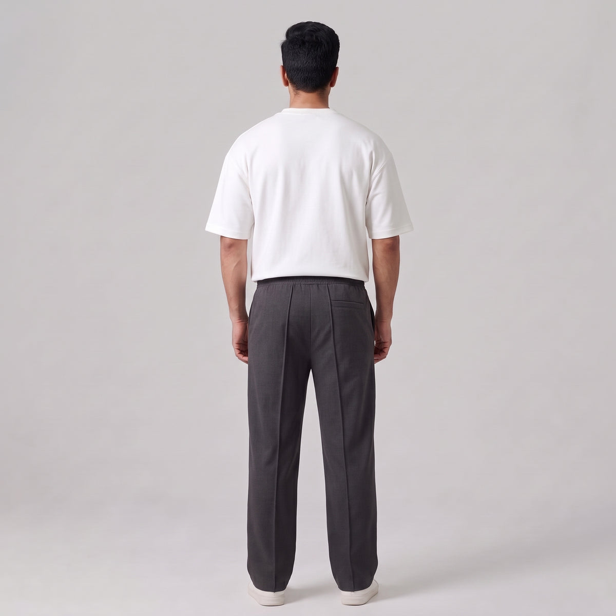 A man viewed from behind wearing a white short-sleeve t-shirt and dark gray baggy  trousers, standing against a light gray studio background l AH Urban Wear