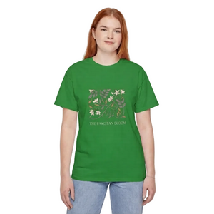 A woman with red hair wearing a green short-sleeve t-shirt with a floral graphic and the text 'THE PAKISTAN BLOOM' on the front, paired with light blue jeans, standing against a solid gray background. l AH Urban Wear Pk