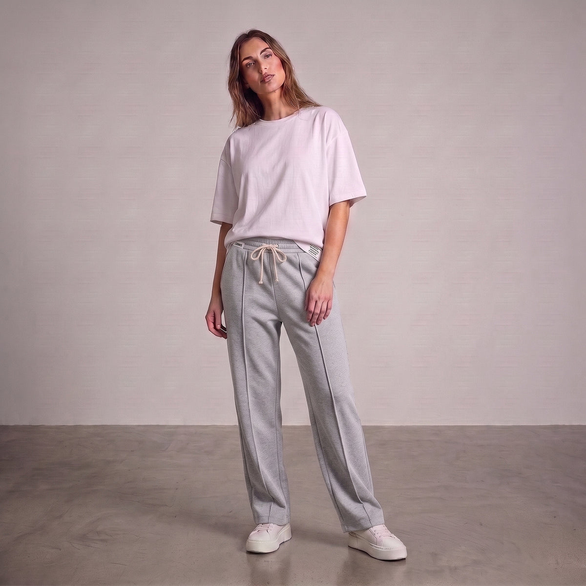 A woman modeling a light pink oversized short-sleeve t-shirt and gray baggy trouser, paired with white sneakers, standing against a neutral studio backdrop l AH Urban Wear