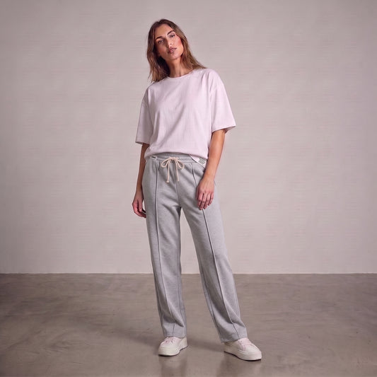 A woman modeling a light pink oversized short-sleeve t-shirt and gray baggy trouser, paired with white sneakers, standing against a neutral studio backdrop l AH Urban Wear