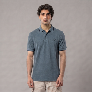 A man wearing a slate gray short-sleeve polo shirt with a small embroidered logo on the chest, paired with light beige pants, standing against a neutral gray studio background | AH Urban Wear PK