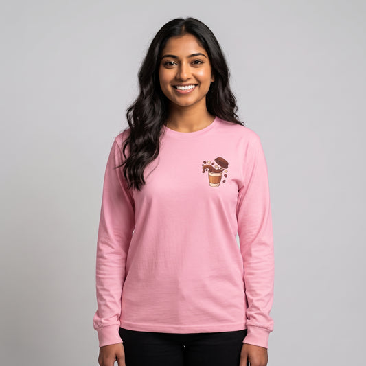 A woman with long dark hair smiling, wearing a light pink long-sleeve crewneck shirt with a small chocolate cup graphic on the left chest, paired with black pants, against a solid gray studio background l AH Urban Wear