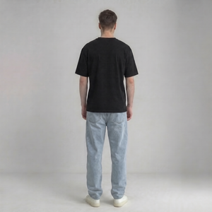 A man viewed from behind wearing a black drop shouder t-shirt, light-wash blue jeans, and white sneakers, standing against a neutral light gray studio background | AH Urban Wear PK