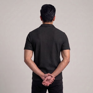 A man viewed from behind wearing a black short-sleeve ribbed knit polo shirt and black pants, standing against a plain light gray studio background with his hands clasped behind his back l AH Urban Wear