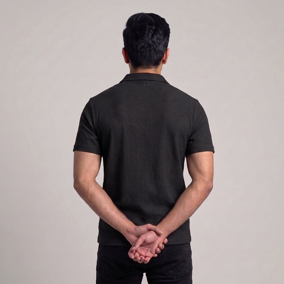 A man viewed from behind wearing a black short-sleeve ribbed knit polo shirt and black pants, standing against a plain light gray studio background with his hands clasped behind his back l AH Urban Wear