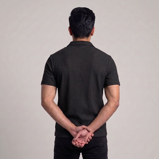 A man viewed from behind wearing a black short-sleeve ribbed knit polo shirt and black pants, standing against a plain light gray studio background with his hands clasped behind his back l AH Urban Wear
