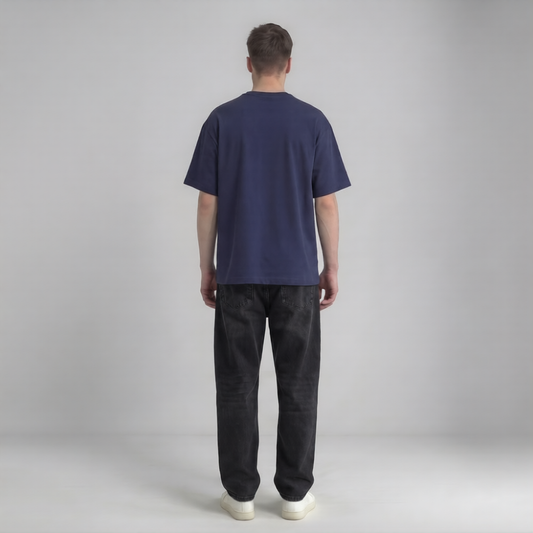 A man viewed from behind wearing a navy blue short-sleeve t-shirt, dark gray washed jeans, and white sneakers, standing against a light gray studio background.