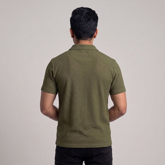 A man viewed from behind wearing a short-sleeve olive green waffle knit polo shirt and black pants, standing against a plain light gray studio background l AH Urban Wear