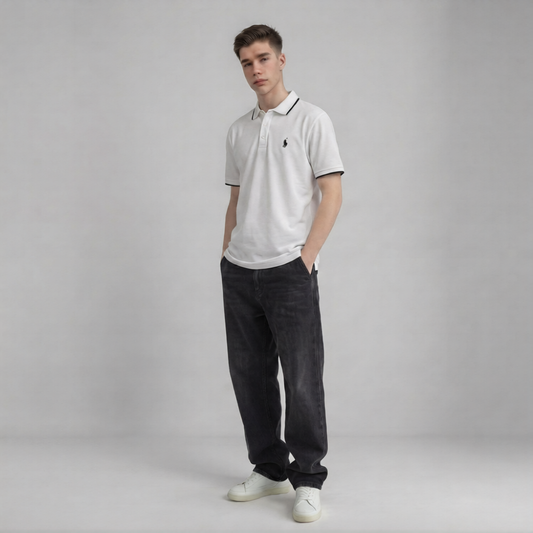 A young man modeling a white short-sleeve polo shirt with a black embroidered chest logo, paired with dark charcoal jeans and white sneakers, standing against a neutral gray studio background l AH Urban Wear