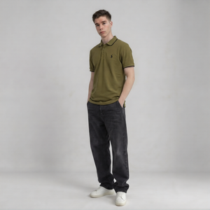 A man modeling an olive green polo shirt with a small embroidered logo on the chest, paired with dark gray jeans and white sneakers, standing against a light gray studio background | AH Urban Wear PK