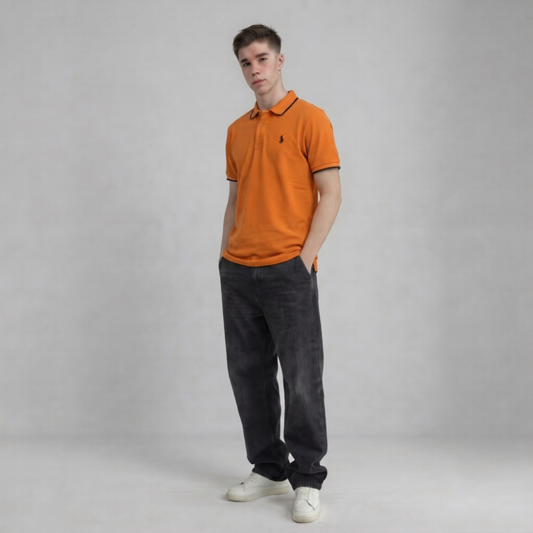 A young man modeling a bright orange short-sleeve polo shirt with black trim and a small embroidered logo on the chest, paired with dark gray jeans and white sneakers, against a neutral gray studio background | AH Urban Wear PK