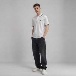 A young man modeling a white short-sleeve polo shirt with a black embroidered chest logo, paired with dark charcoal jeans and white sneakers, standing against a neutral gray studio background | AH Urban Wear PK