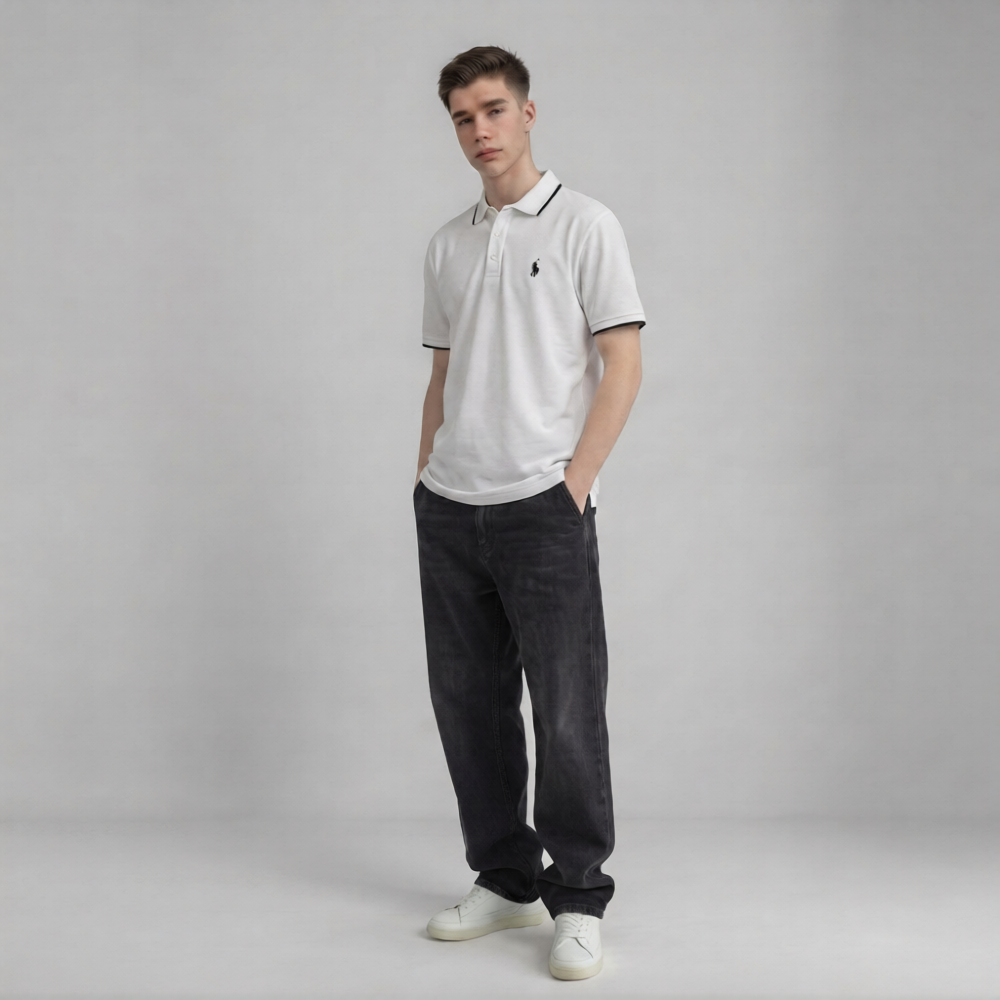 A young man modeling a white short-sleeve polo shirt with a black embroidered chest logo, paired with dark charcoal jeans and white sneakers, standing against a neutral gray studio background | AH Urban Wear PK