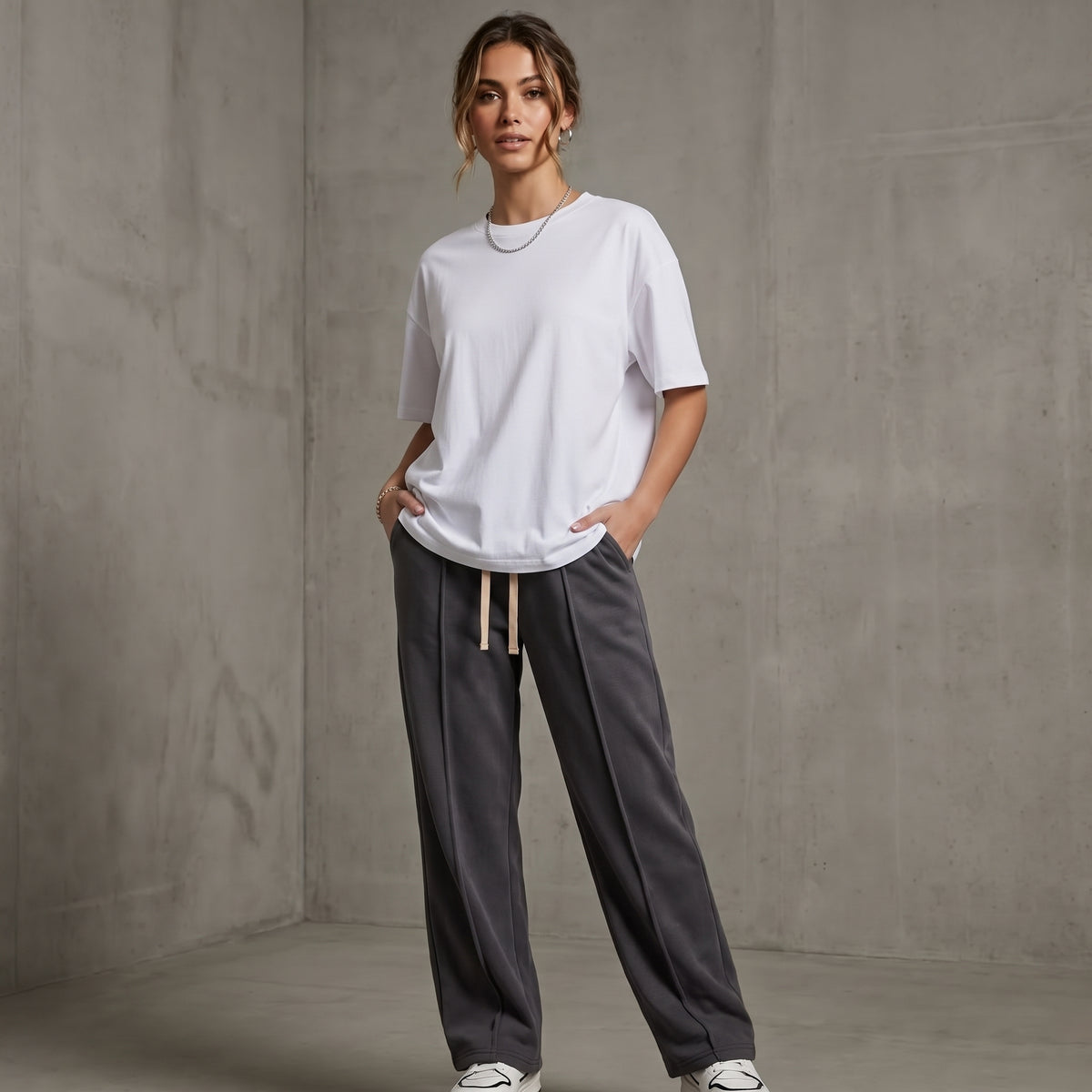 A woman modeling a white short-sleeve crewneck t-shirt and dark gray baggy trouser with a beige drawstring, standing against a textured gray concrete wall l AH Urban Wear