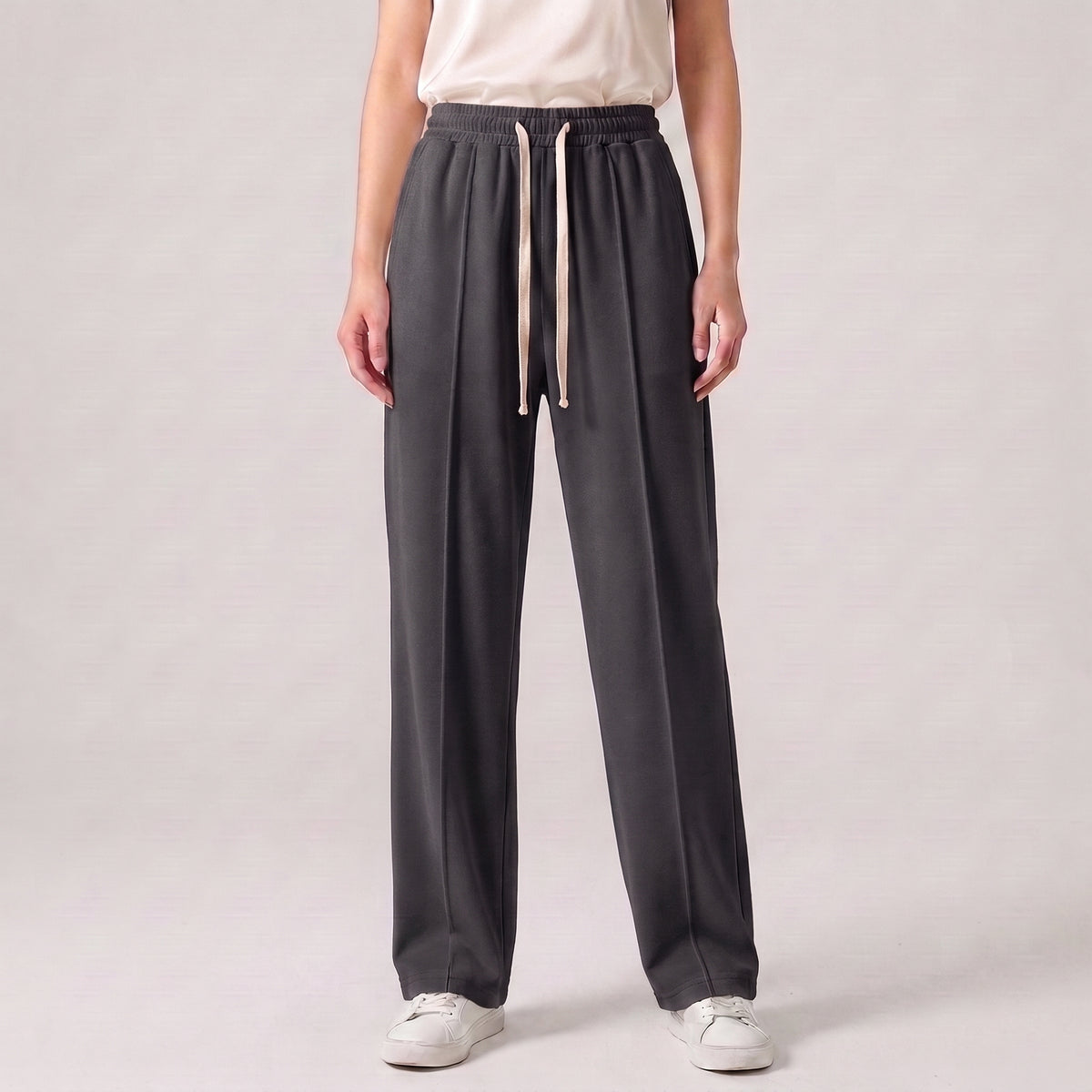 A person wearing dark charcoal gray wide-leg drawstring pants, a light beige top, and white sneakers, standing against a light gray studio background l AH Urban Wear