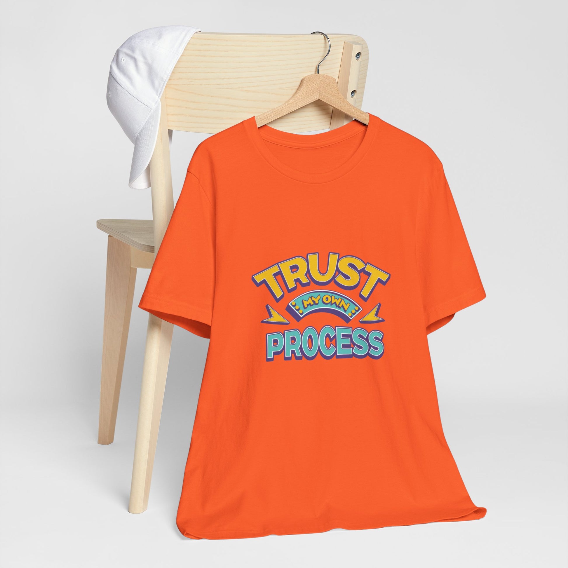 An orange short-sleeve t-shirt with a colorful graphic reading 'TRUST MY OWN PROCESS' hanging on a wooden hanger, displayed in front of a light wood chair with a white shirt draped over it, against a light gray background | AH Urban Wear PK
