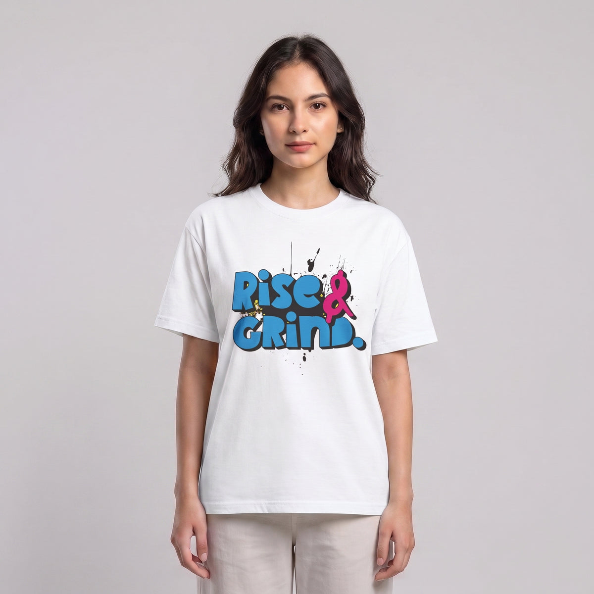 A woman modeling a white short-sleeve t-shirt with a colorful graphic on the chest that reads 'Rise & Grind.' in blue and pink graffiti-style lettering, set against a plain light gray studio background l AH Urban Wear