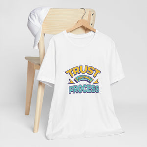 A white short-sleeve t-shirt on a wooden hanger, styled in front of a light wood chair with a white cap draped over the back. The shirt features a colorful retro-style graphic on the chest reading 'TRUST MY OWN PROCESS"  | AH Urban Wear PK