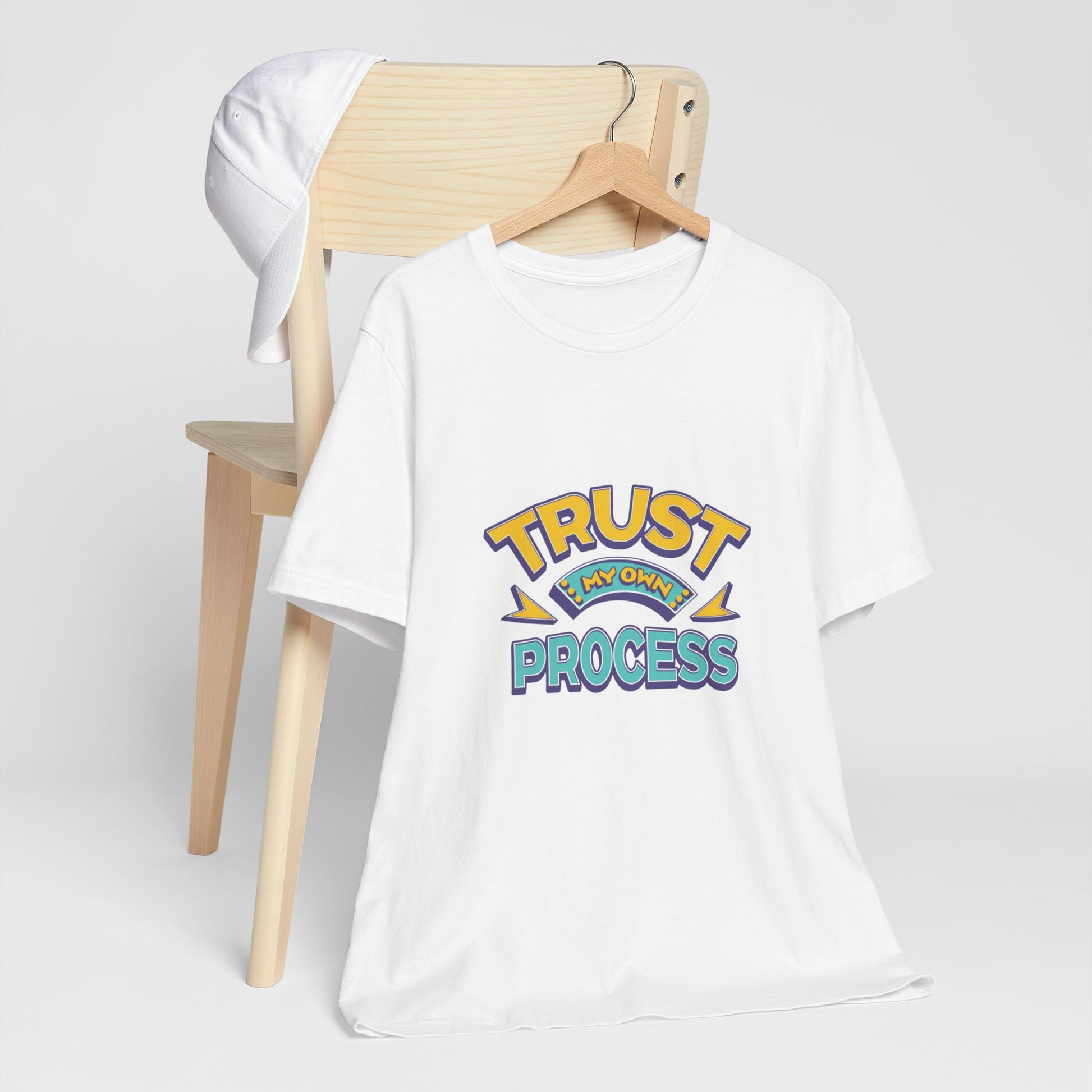 A white short-sleeve t-shirt on a wooden hanger, styled in front of a light wood chair with a white cap draped over the back. The shirt features a colorful retro-style graphic on the chest reading 'TRUST MY OWN PROCESS"  | AH Urban Wear PK