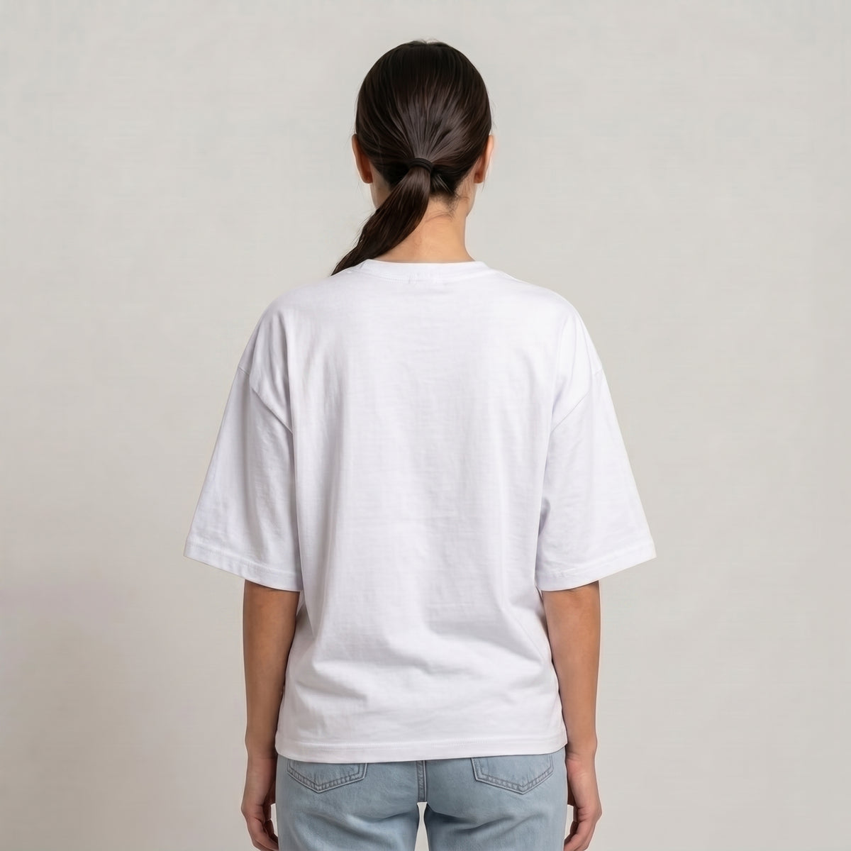 A woman viewed from behind wearing a plain white drop shoulder t-shirt and light blue jeans, standing against a neutral light gray studio background | AH Urban Wear 