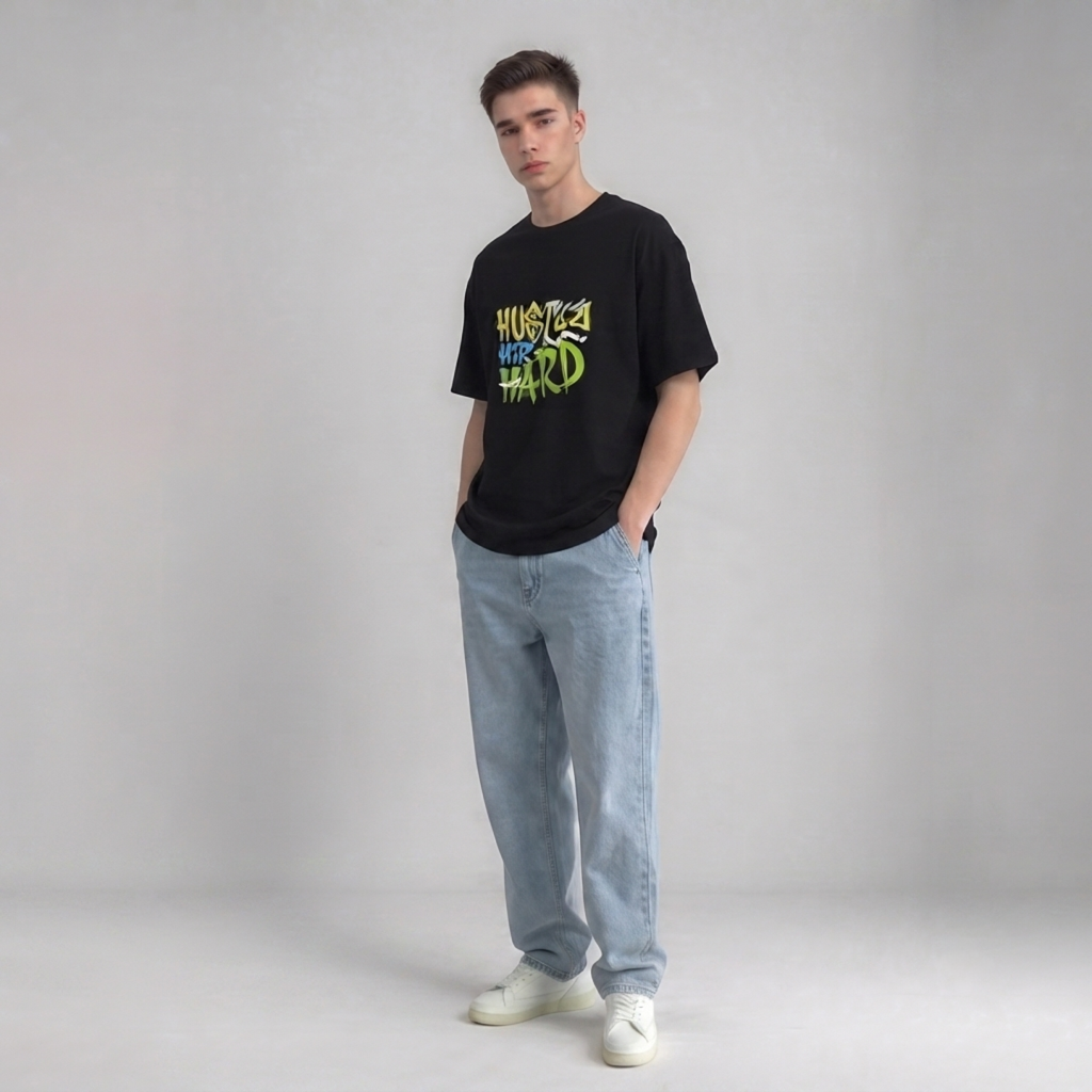 A young man modeling a black graphic drop shoulder t-shirt with 'HUSTLE YOUR HARD' printed in green and yellow, paired with light blue jeans and white sneakers, against a neutral gray studio background | AH Urban Wear PK