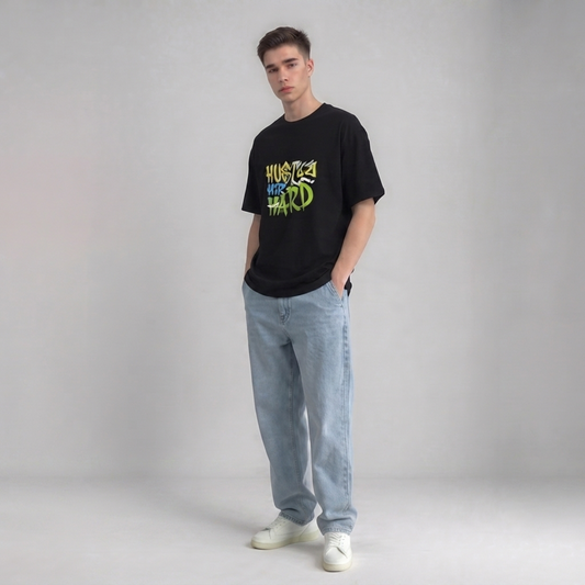 A young man modeling a black graphic drop shoulder t-shirt with 'HUSTLE YOUR HARD' printed in green and yellow, paired with light blue jeans and white sneakers, against a neutral gray studio background | AH Urban Wear PK
