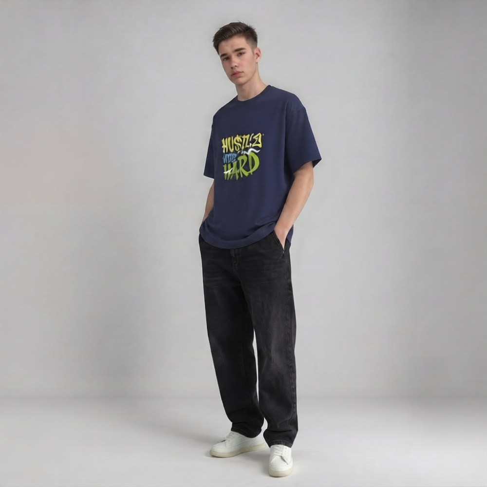 A young man modeling a navy blue drop shoulder t-shirt with a green and yellow graphic that reads 'HUSTLE HIT HARD', paired with dark jeans and white sneakers, against a light gray studio background | AH Urban Wear PK